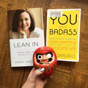 A photo of books and a one-eyed daruma figure. The books are Lean In by Sheryl Sandberg and You Are a Badass by Jen Sincero.